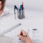 Profissional estudando desenho técnico mecânico com projetos detalhados em papel e computador.