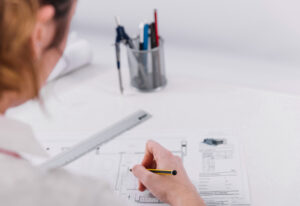 Profissional estudando desenho técnico mecânico com projetos detalhados em papel e computador.